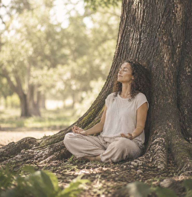 Mujer meditando apoyada en un árbol en el bosque, en actitud de presencia y autoconocimiento